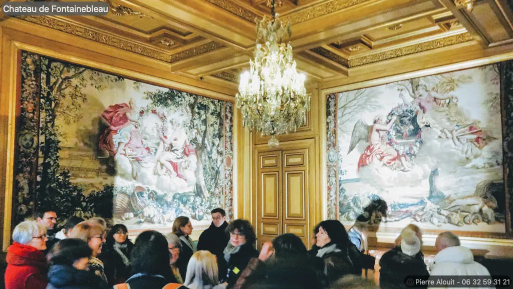Une photo d'une salle du Château de Fontainebleau, où un groupe de personnes participe à une visite guidée. La pièce est ornée de deux grandes tapisseries murales représentant des scènes mythologiques ou historiques avec des personnages en costume d'époque. Un lustre en cristal illumine la pièce, et le plafond est décoré de caissons dorés. Des portes en bois double battant sont également visibles sur le côté de la pièce.