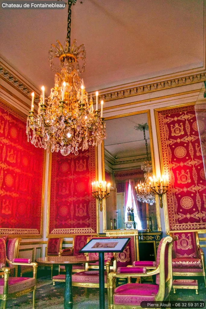 Une photo d'une salle au Château de Fontainebleau avec des murs habillés de tentures rouges ornées d'éléments dorés et du symbole de l'abeille, caractéristique du style impérial. Un lustre en cristal illumine la pièce, suspendu à un plafond orné de moulures dorées. Le mobilier comprend des chaises dorées à l'assise et au dossier recouverts de velours rouge, et une cheminée en marbre est visible en arrière-plan. Un panneau d'information est placé devant une table, suggérant un espace de visite guidée dans le château.